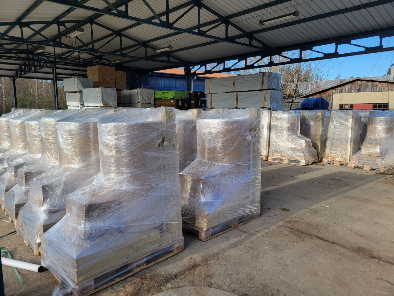Stacks of palletized greenhouses under a metal roof with clear plastic wrapping.