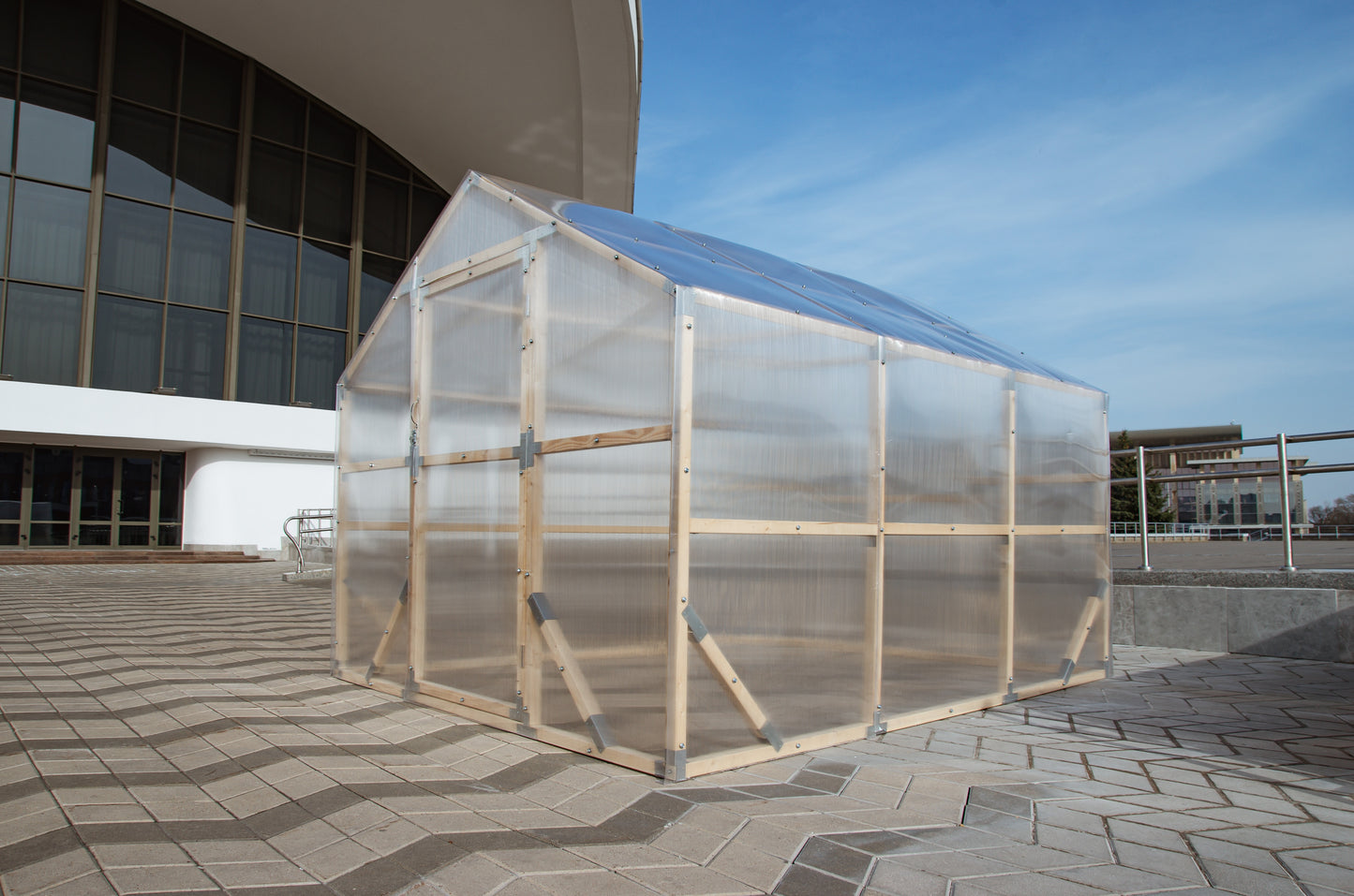 Transparent greenhouse structure on a paved area with a building in the background