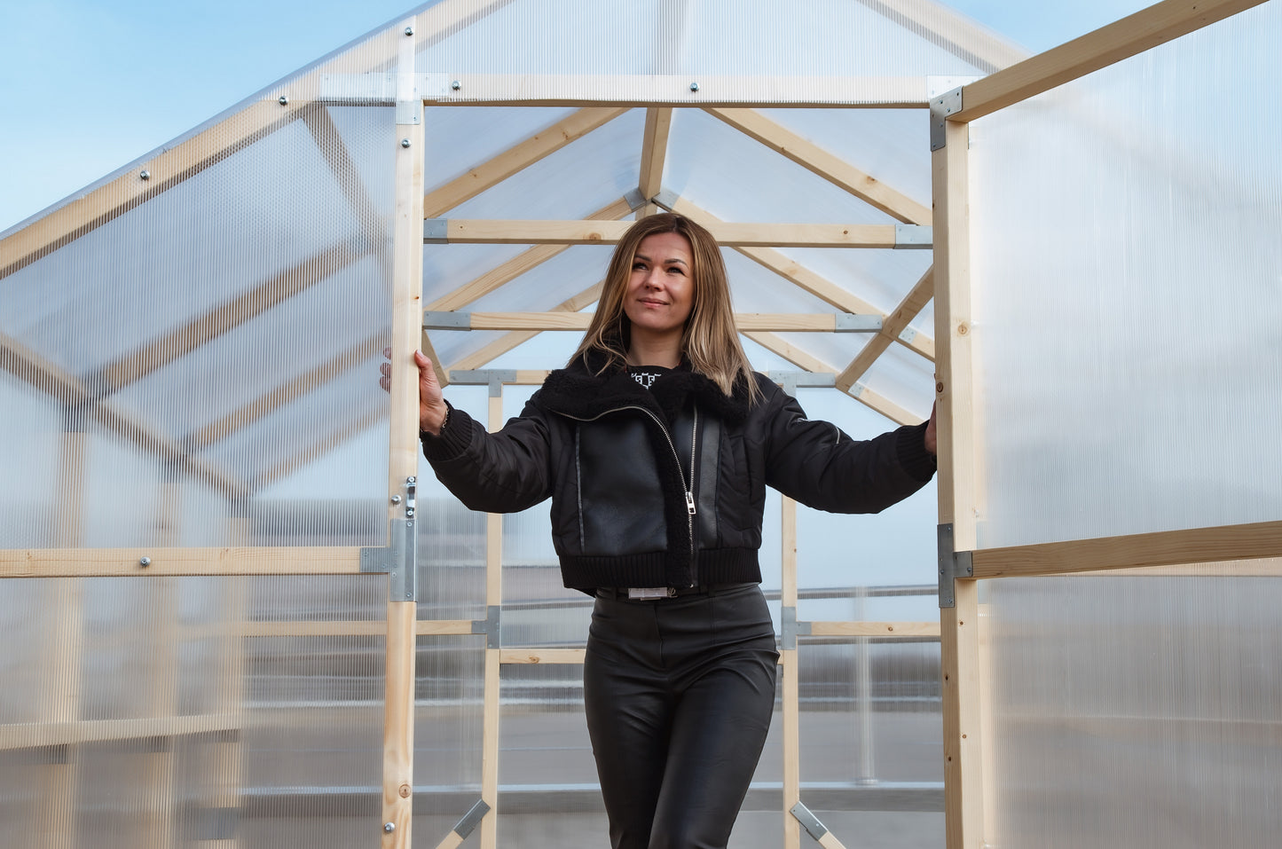 Person standing inside a transparent greenhouse with wooden frames.