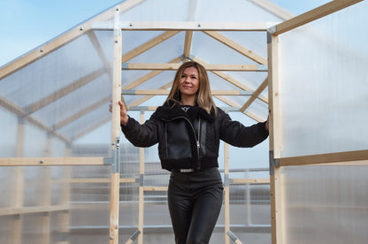 Person standing inside a transparent greenhouse with wooden frames.